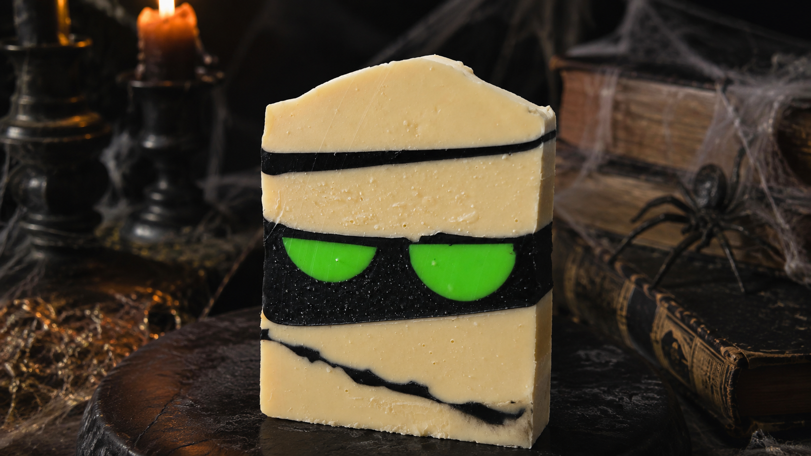 Savon artisanal Les Mels façon momie, bandelettes crème et noires avec deux yeux verts, posé sur un décor de vieux grimoires et toiles d'araignée